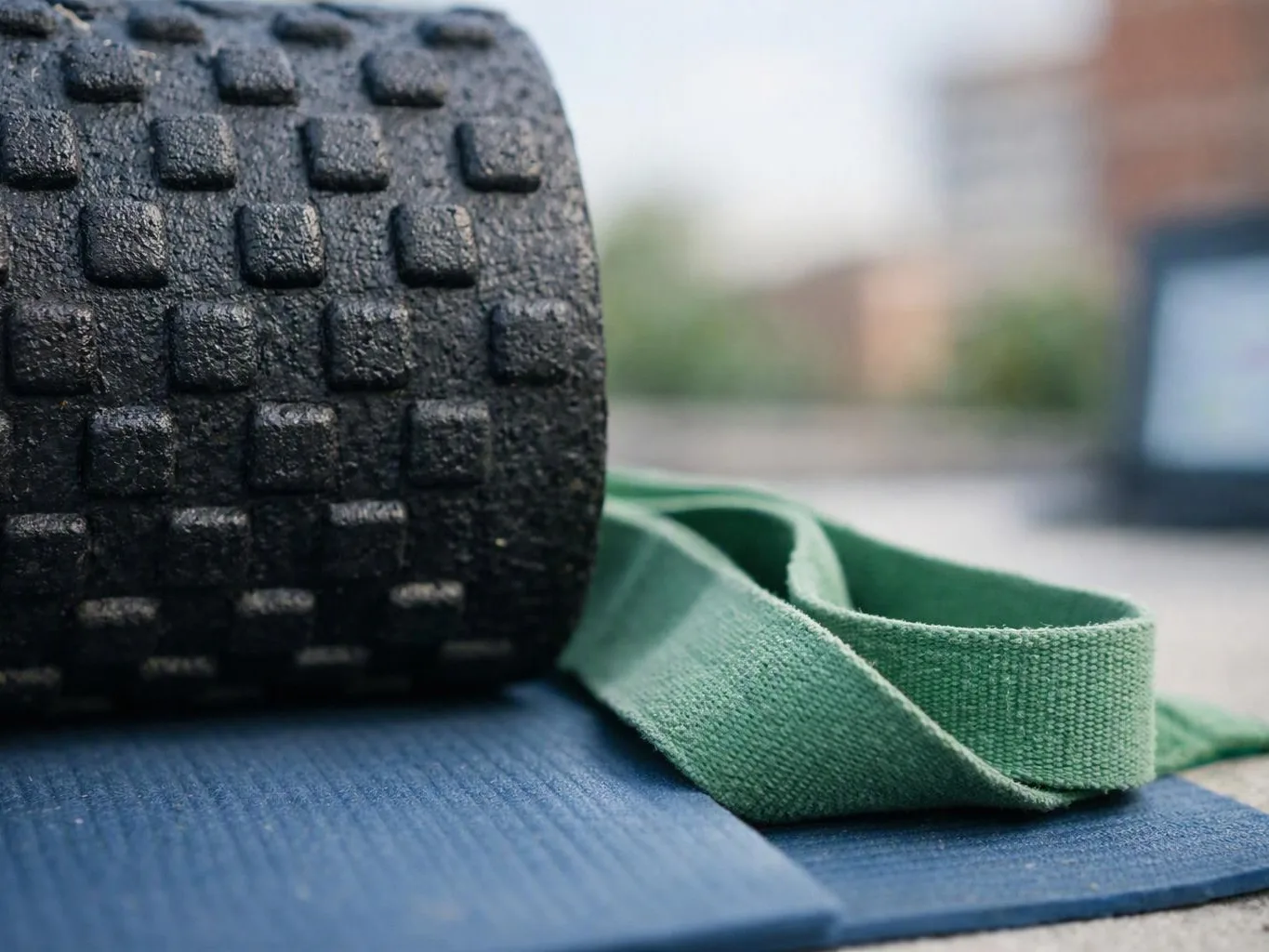 A black textured foam roller rests on a blue yoga mat beside a green strap outdoors