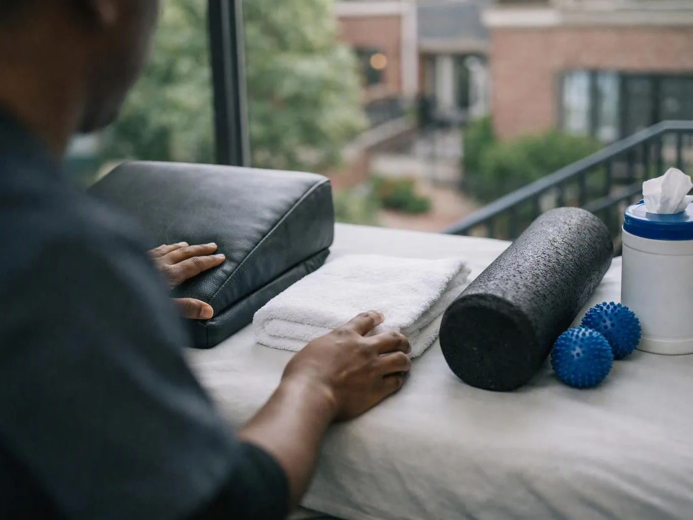 A person arranges towels, foam rollers, massage balls, and a tissue container on a table by a window